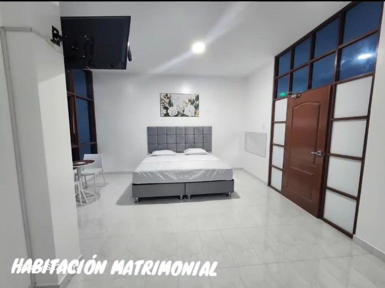 Modern minimalist bedroom with gray upholstered bed, white flooring, and floor-to-ceiling glass doors, labeled Habitacion Matrimonial
