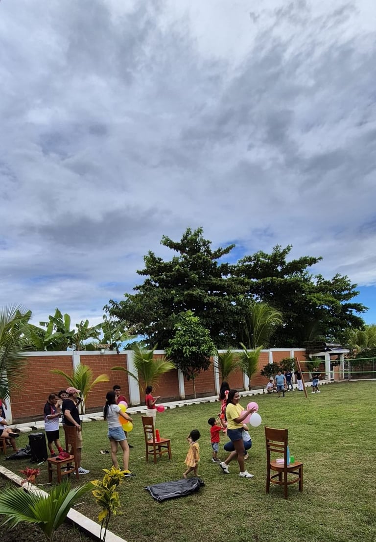 Children and adults playing outdoors on a grassy field with a brick building and palm trees under a cloudy sky
