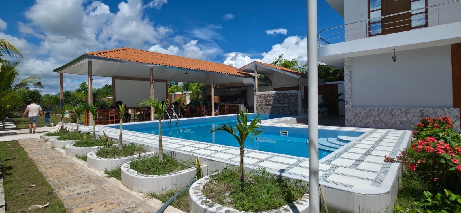 Modern residential house with rectangular swimming pool, white patio, and covered pergola surrounded by landscaped garden under blue sky