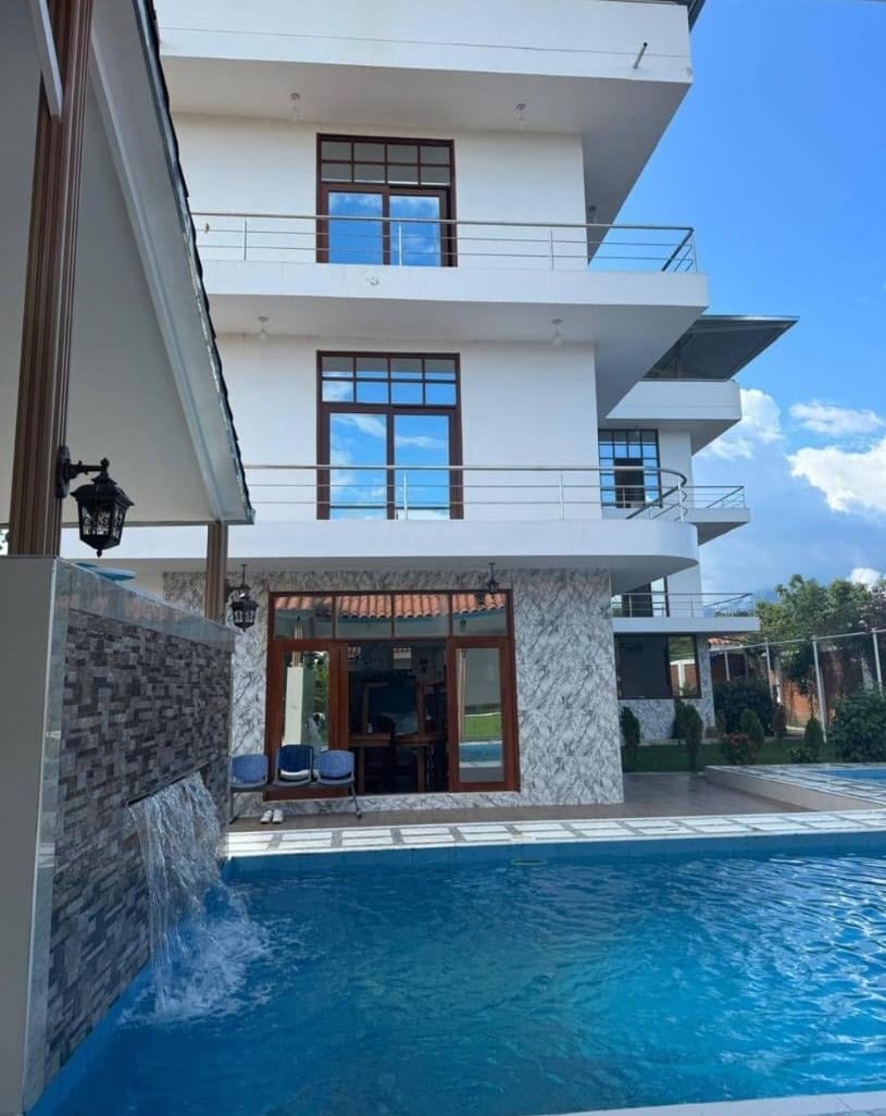 Modern white multi-story residential building with pool, featuring metal railings, wooden entrance doors, and stone accents under blue sky