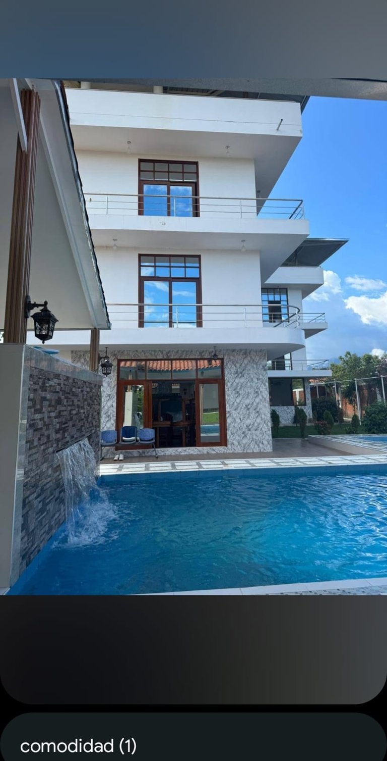 Modern white multi-story residential building with blue-framed windows, metal railings, and a swimming pool in the foreground under clear sky